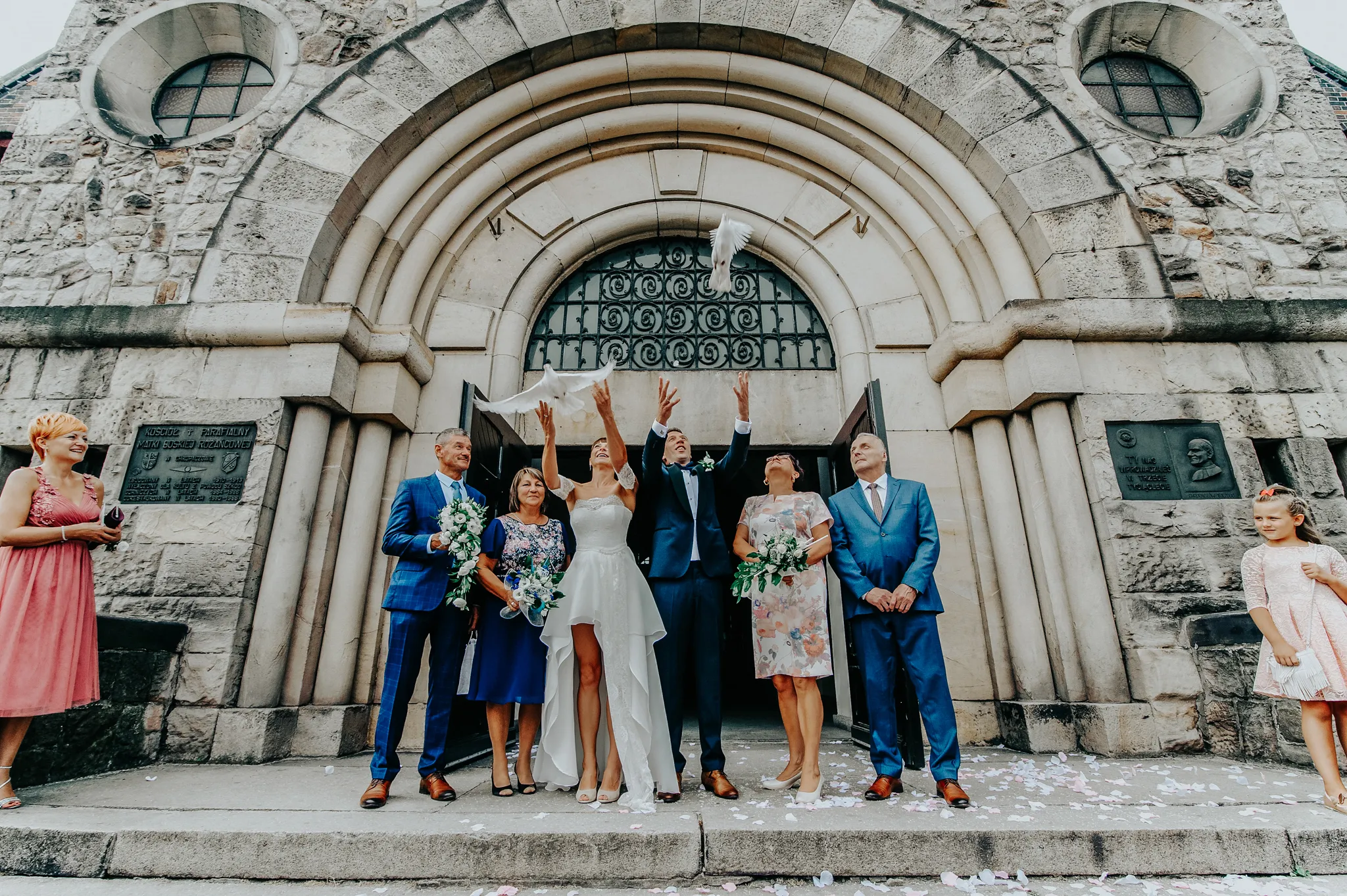 mloda para stoi przed kosciolem w zabrzu wypuszczajac z rak golebie w otoczeniu rodzicow podziwiajacych ulatujace ptaki . fotograf slubny katowice